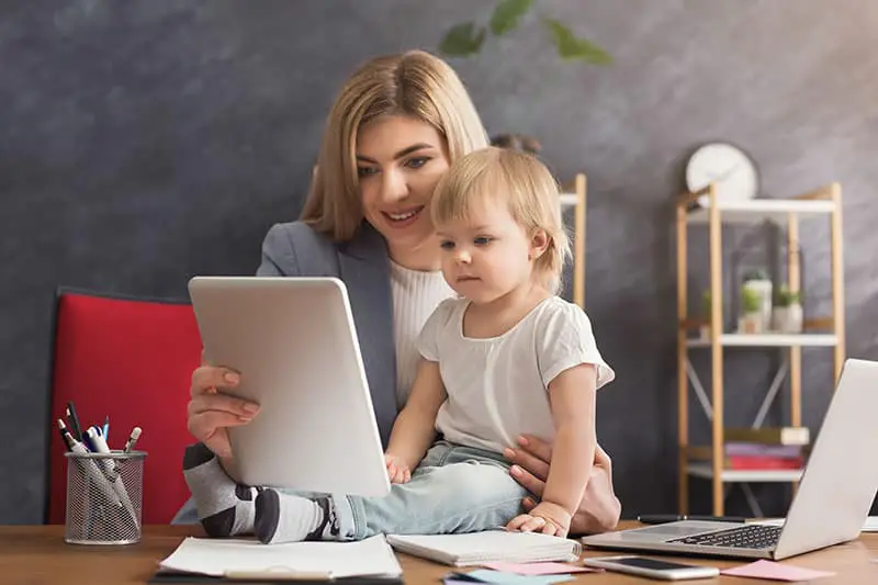 Happy beautiful business mom using digital tablet while spending time with her cute baby. Business, motherhood, multitasking and family concept.