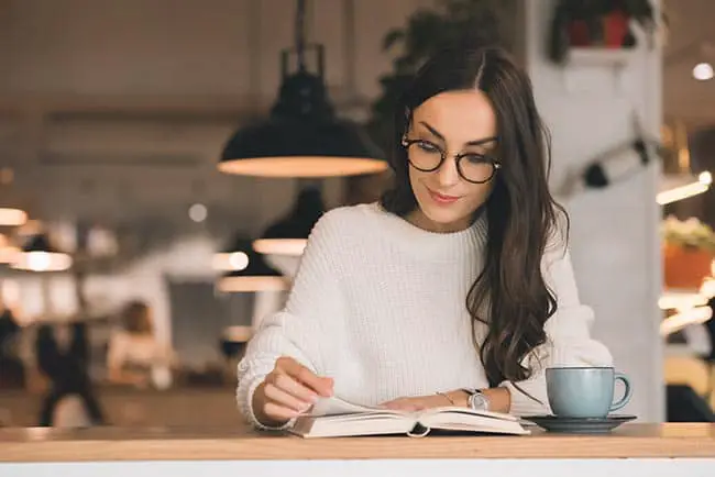 Attractive young woman in eyeglasses reading book at table with coffee cup in cafe