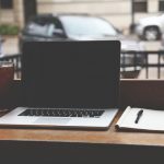 MacBook Pro on a wooden table with iced coffee and notebook in a casual coffee shop setting.